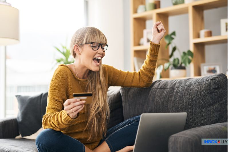 Woman sitting on a couch, holding a credit card and raising her fist in excitement while looking at a laptop—celebrating mastering Bulk Email Practices for 2024.