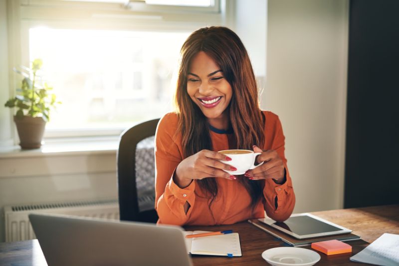 A smiling woman drinking coffee and reading emails on a laptop.