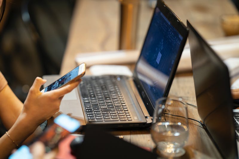 A woman is using her phone while sitting at a table with a laptop.