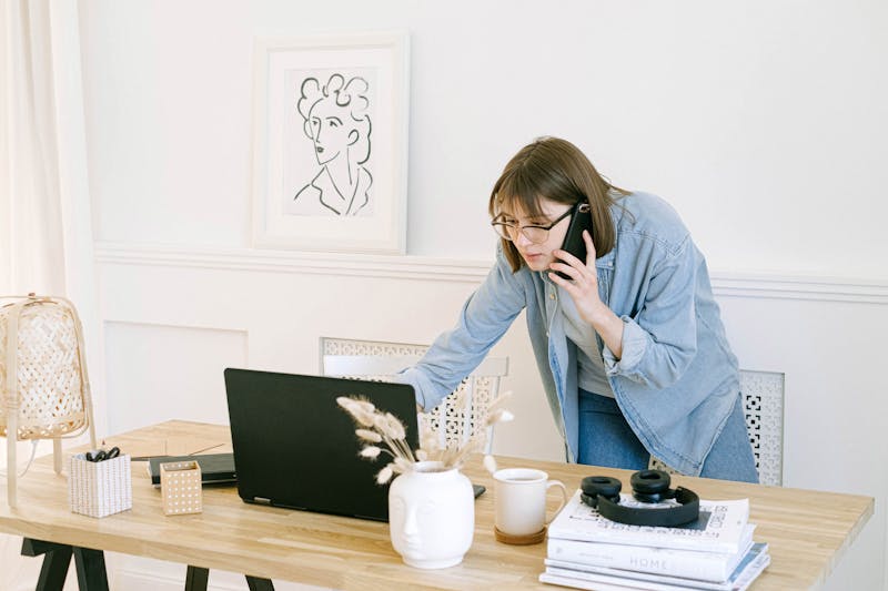 Mulher multitarefa: receber uma ligação telefônica enquanto trabalha em um laptop, enfrentando o desafio de gerenciar vários registros SPF para um domínio.