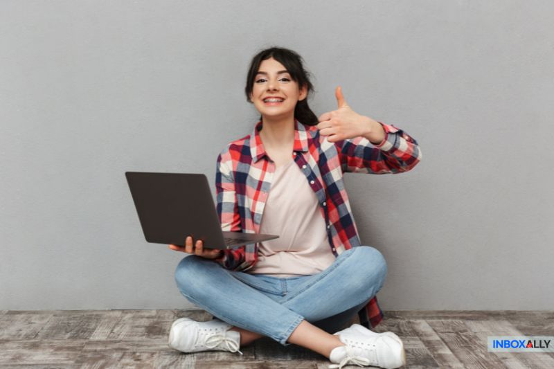Sitting cross-legged on the floor in a plaid shirt and jeans, the person confidently gives a thumbs-up while holding their laptop, ready to tackle tasks like spam trap detection against a plain gray backdrop.