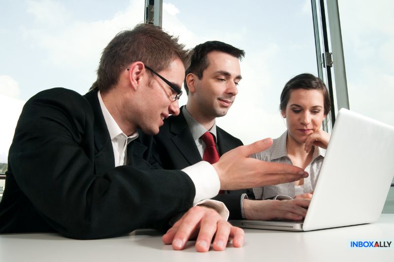 Three people in formal attire sit at a table, engaged with a laptop. One person gestures while speaking passionately about Yahoo blocking their emails, another listens attentively, and the third looks intently at the screen.