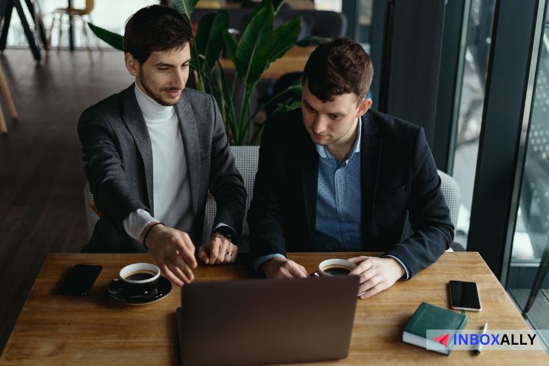 Two men in business attire sit at a wooden table, engaging in a discussion about the best LinkedIn outreach strategies while looking at a laptop. 