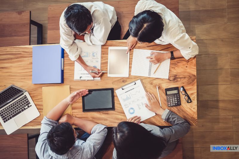 Four people sit around a wooden table with laptops, tablets, papers, a calculator, and a notebook, engaged in a lively discussion on growth hack strategies and reputation gamble techniques.