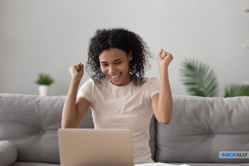 A person sits on a couch with a laptop, raising their fists in excitement after successfully optimizing their email marketing strategy.