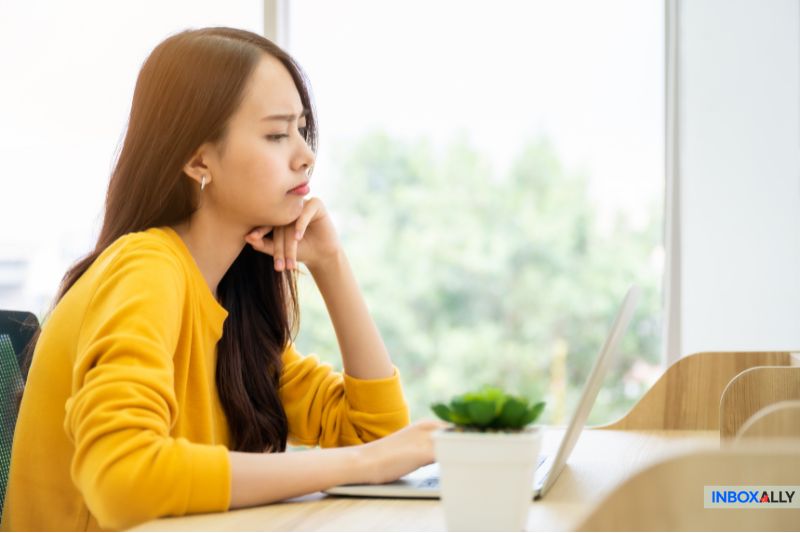 A person in a yellow sweater is sitting at a desk, looking at a laptop screen with a thoughtful expression, pondering over reputation management.