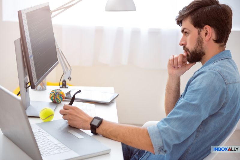 A man works on a laptop at a desk, analyzing email metadata with focused intent. Nearby, a computer monitor, tablet, and desk toys create an engaging workspace that bolsters his concentration on improving deliverability.