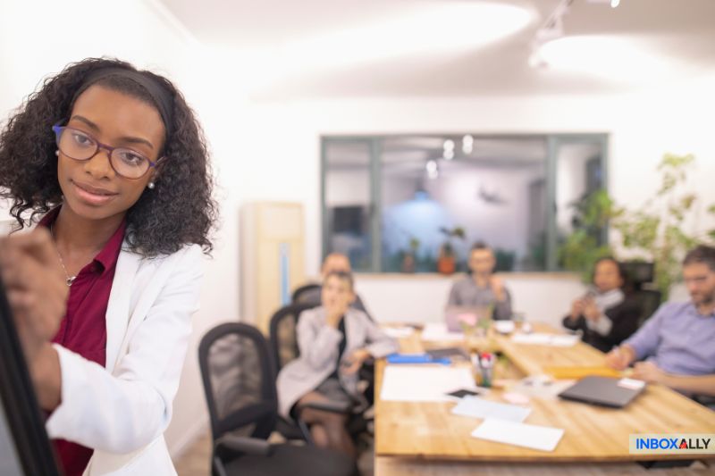 Uma mulher em frente a um quadro branco em uma sala de reuniões, ilustrando a importância da taxa de cliques, enquanto várias pessoas sentadas em uma mesa ao fundo participam de uma discussão animada sobre essa métrica crucial.
