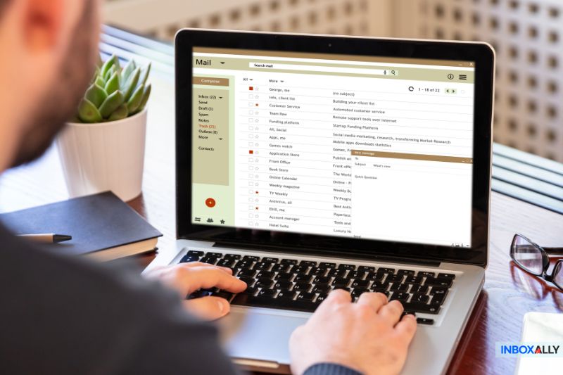 A person is checking email on a laptop, displaying an inbox filled with numerous unread messages from subscribers. A notebook and plant rest on the wooden table.