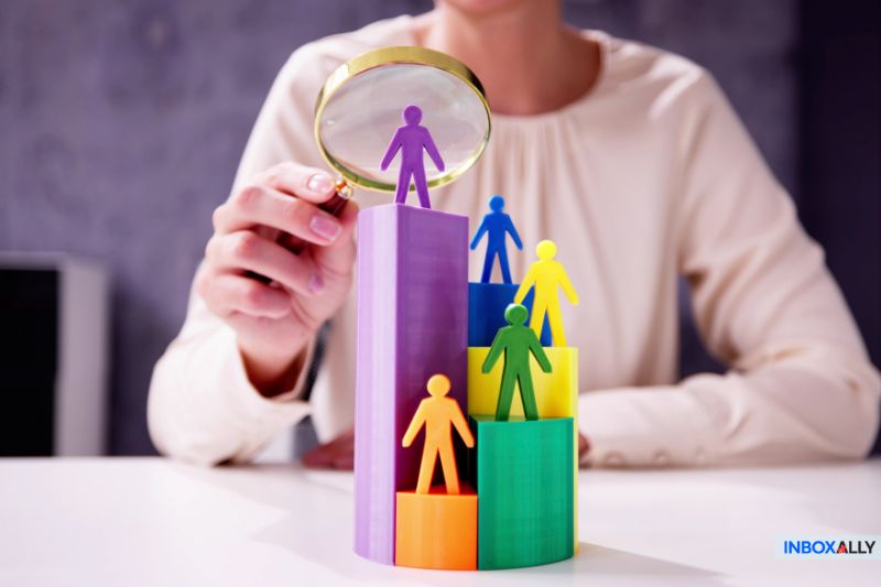 A person holds a magnifying glass over colorful humanoid figures positioned on a bar graph, symbolizing the intricate process of understanding subscribers in email lead generation.
