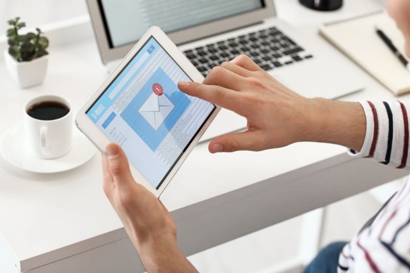 Uma pessoa está efetivamente lendo um e-mail em um tablet em uma mesa, cercada por um laptop, uma xícara de café e um notebook. Parece que eles estão se preparando para enfrentar os desafios do mundo digital em ritmo acelerado de 2025.