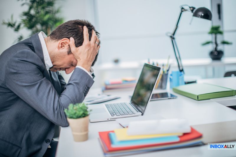 A man sits at his desk with his head in his hands, looking frustrated by a 550 5.1.1 Error on his laptop screen. A stack of folders, potted plant, and desk lamp surround him as he contemplates steps to avoid future errors.