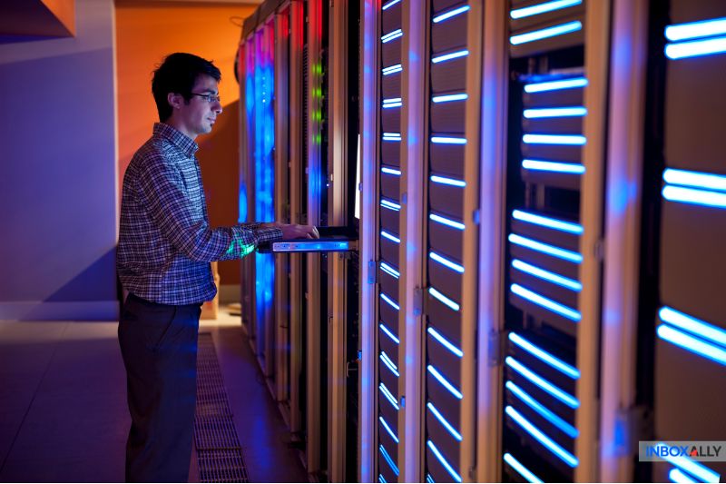 In a server room, blue and green lights flicker as a person operates a laptop, meticulously avoiding any user unknown errors that could disrupt the process.