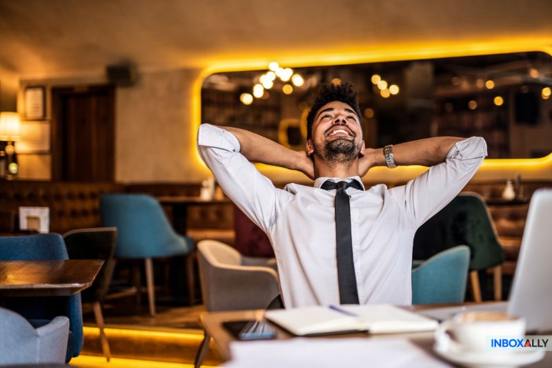 A person in a tie leans back at a café, grinning with hands behind their head, successfully avoiding the "User Unknown" message on their laptop. A notebook sits nearby as they enjoy this moment of peace amidst the usual tech hiccups.
