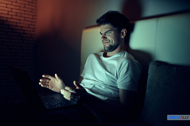In a dimly lit room, a man in a white t-shirt looks frustrated while using his laptop, overwhelmed by the intricacies of AI email personalization.