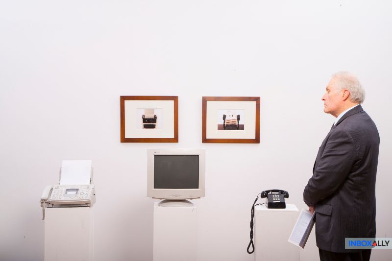 A man in a suit observes two framed photographs above a once-buzzing fax machine, CRT monitor, and rotary phone on pedestals—a nostalgic display juxtaposed with the modern precision of an email spam filter in today's hosted email solutions.