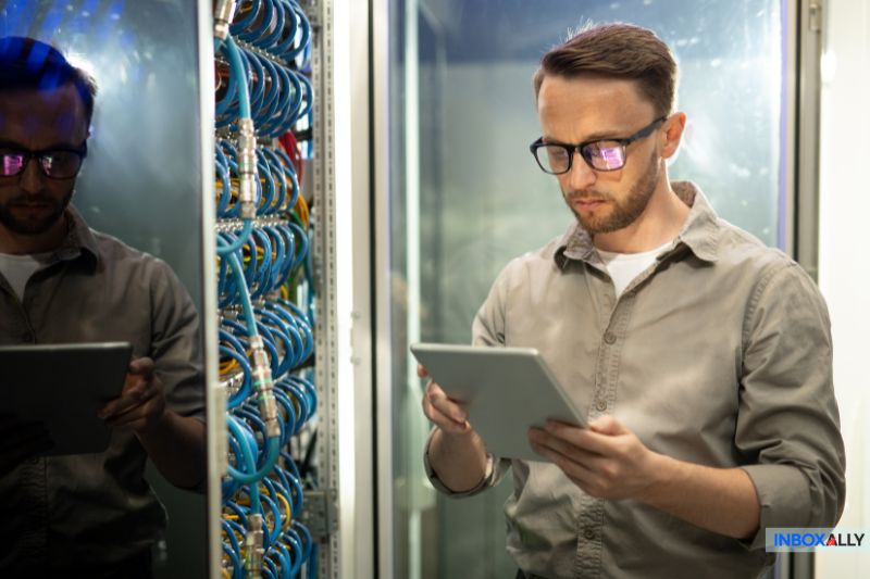 In a server room, a man wearing glasses focuses on his tablet, diligently checking SMTP configuration details. Wires and equipment fill the background, creating an atmosphere of high-tech precision.