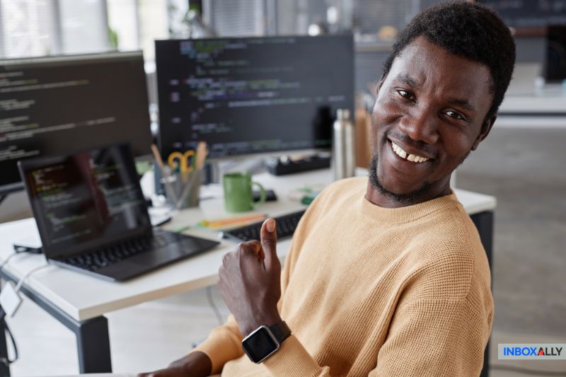 A person smiles and gives a thumbs-up while sitting at a desk with a laptop showing code, possibly setting up SMTP settings for Hotmail in an office filled with multiple desks and monitors.