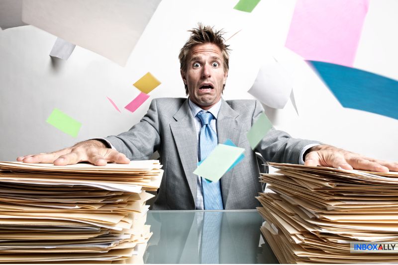 A man in a suit looks overwhelmed as colorful papers fly around him, surrounded by two large stacks of files on a desk, reminiscent of his chaotic inbox before email syncing simplified his life.