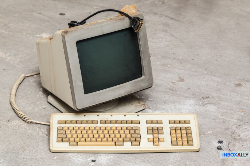 An old, yellowed CRT monitor with a detached cable sits on the floor next to a vintage keyboard, harking back to a time when email syncing was just taking root with protocols like POP3 and IMAP.