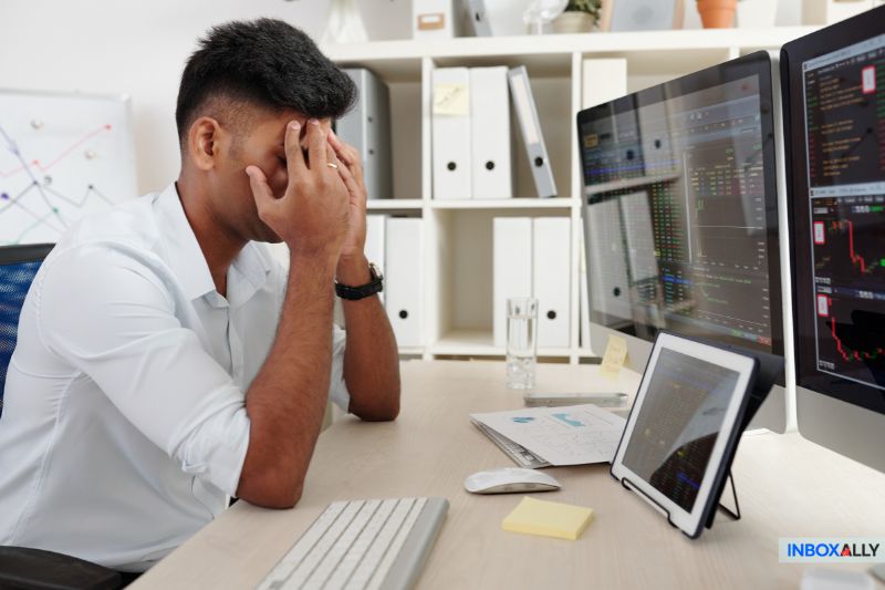 A man sits at his desk, hands on his face, looking stressed as multiple computer screens display financial data. The inboxally logo is visible, indicating issues with emails syncing via IMAP.