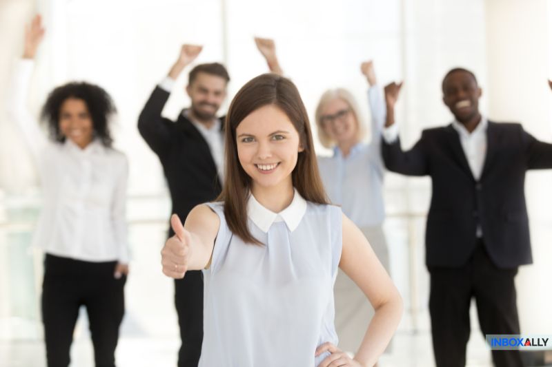 A woman gives a thumbs up, standing in front of four people with raised arms, all smiling, celebrating their successful email syncing in a bright office setting.