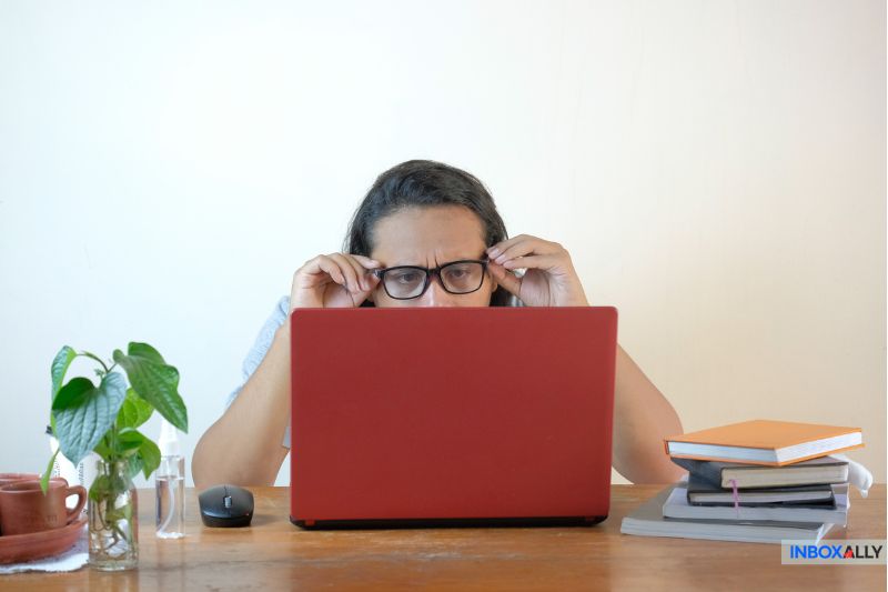 A person adjusts their glasses while looking worriedly at a red laptop, seated at a table with a plant, a cup, notebooks, and a pen.
