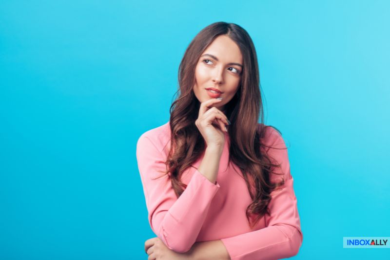 A woman in a pink top stands against a blue background, her hand on her chin, appearing thoughtful yet slightly worried.