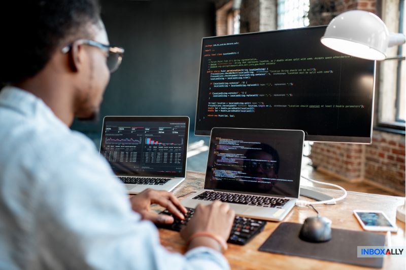 A person types on a keyboard at a desk with two laptops displaying code and data, ensuring email security through detailed DMARC record analysis. A desk lamp is next to them in a well-lit room.