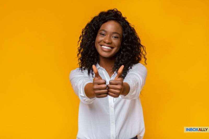 A person with curly hair smiles and gives two thumbs up while wearing a white shirt, exuding confidence and security against a vibrant yellow background.