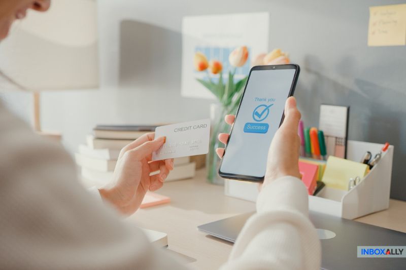 A person grins while holding a credit card and a smartphone showing a "Success" message, seated at a desk cluttered with a laptop and office supplies. They've avoided spam and the hassle of fake order confirmation emails with their savvy online shopping skills.