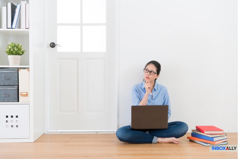 Uma mulher está sentada no chão, pensativa, com seu laptop, explorando as principais opções de soluções de e-mail. Os livros estão empilhados ao seu lado enquanto ela se encosta na parede branca em frente a uma porta, mergulhando em discussões sobre as melhores opções de Sendgrid disponíveis.