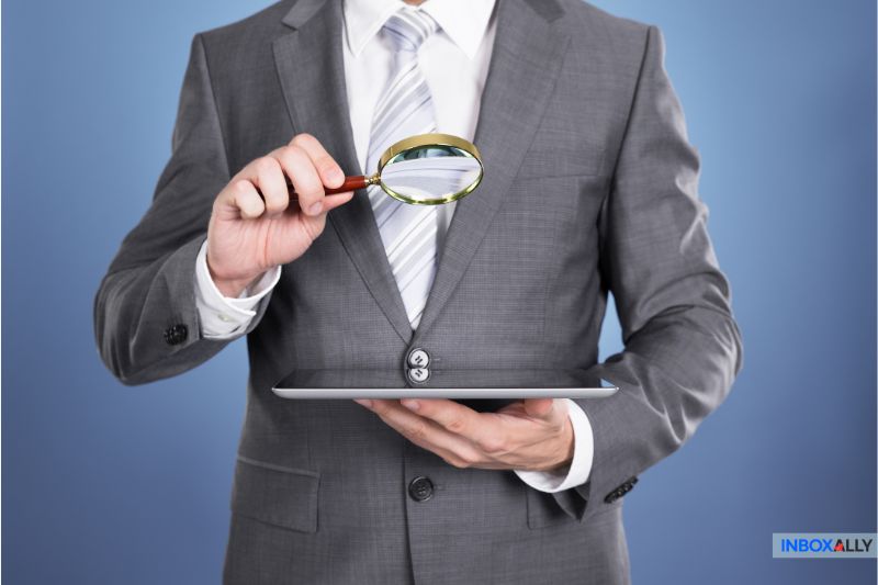 A man in a suit, exploring AWeber Alternatives for 2025, holds a magnifying glass over a tablet against a blue background.