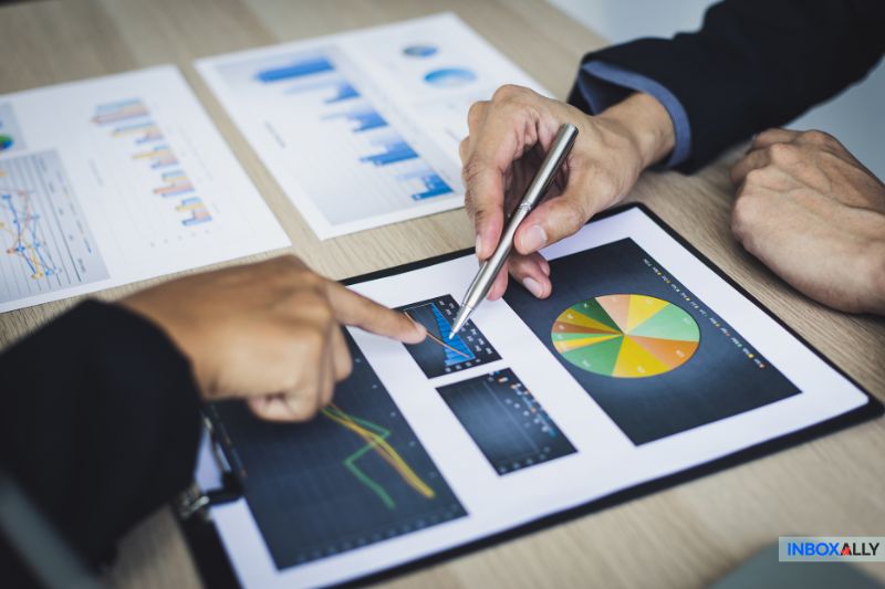 Two people are analyzing charts on a clipboard and a tablet, discussing strategies for 2025. Several graphs and email marketing plans are also visible on papers scattered across the desk.