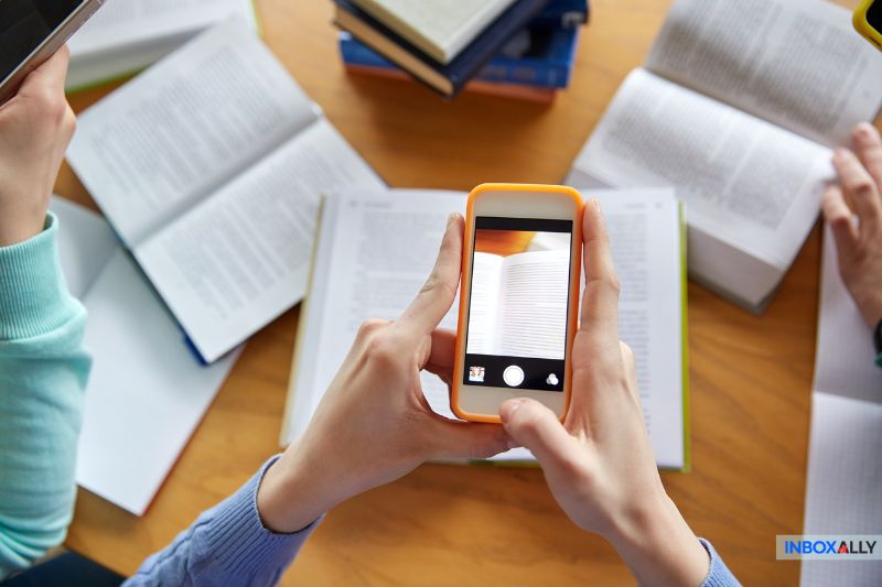 A person takes a photo of an open book with a smartphone while other open books and a stack of books—along with a note about GoDaddy Email Settings—are on the table.