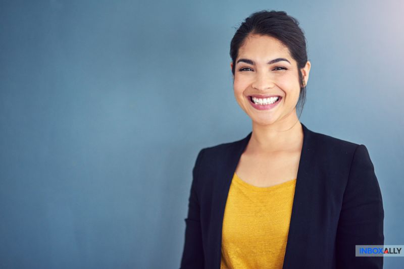 A woman wearing a black blazer and yellow top smiles in front of a plain blue background, ready to help you navigate GoDaddy Email Settings.