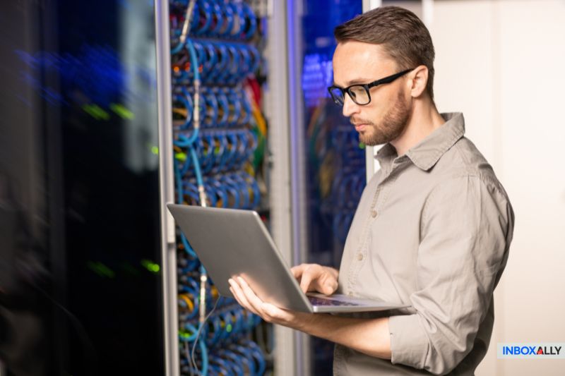 A man using a laptop stands in a server room with network cables and server racks visible, possibly reviewing GoDaddy SMTP settings. The INBOXALLY logo appears in the bottom right corner.