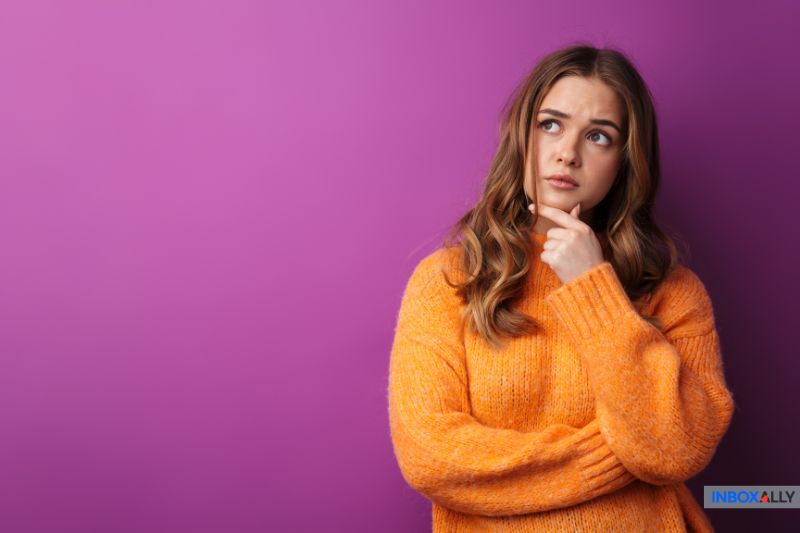 A woman in an orange sweater stands against a purple background, looking up with a thoughtful expression and her hand on her chin, as if pondering GoDaddy SMTP settings for email configuration.