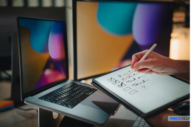 Uma pessoa usa uma caneta stylus para marcar itens em uma lista de verificação digital sobre autenticação de e-mail da GoDaddy, exibida em um tablet, com um laptop e um monitor em segundo plano.
