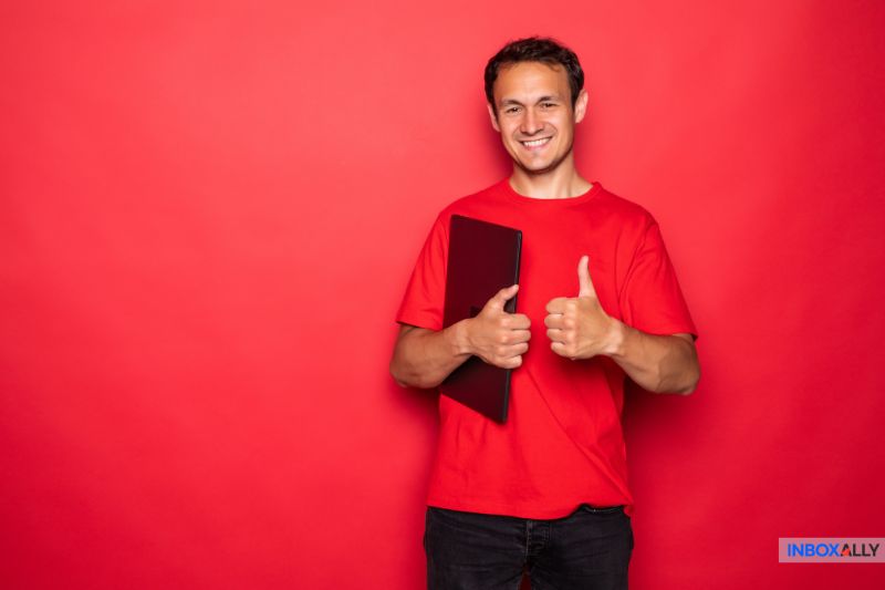 Um homem de camisa vermelha segura um laptop fechado e faz um gesto de polegar para cima enquanto está de pé contra um fundo vermelho, comemorando a autenticação bem-sucedida de e-mail e a configuração de SPF da GoDaddy.