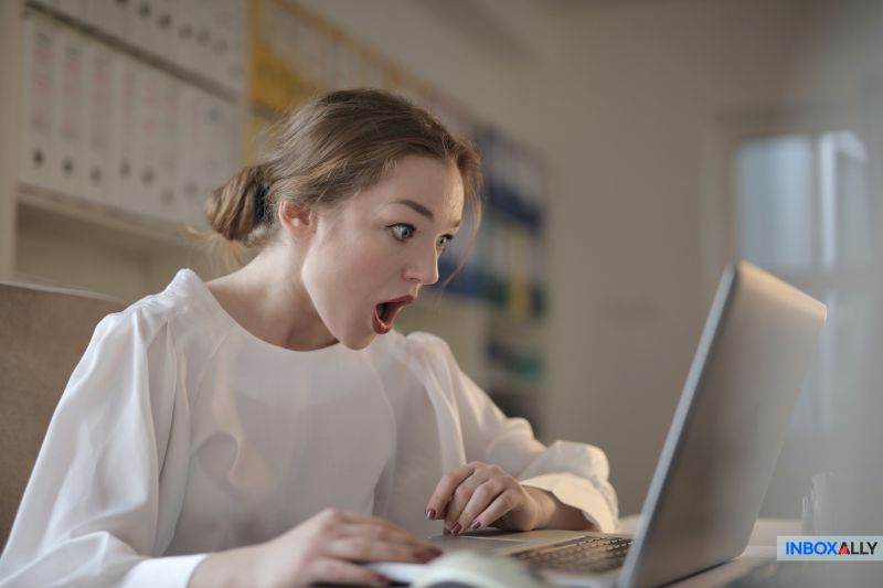 Uma mulher com expressão de surpresa olha para a tela de um laptop enquanto está sentada em uma mesa, reagindo a uma notificação de erro de SPF. O logotipo da InboxAlly está visível no canto inferior direito, destacando as preocupações com a capacidade de entrega de e-mails.