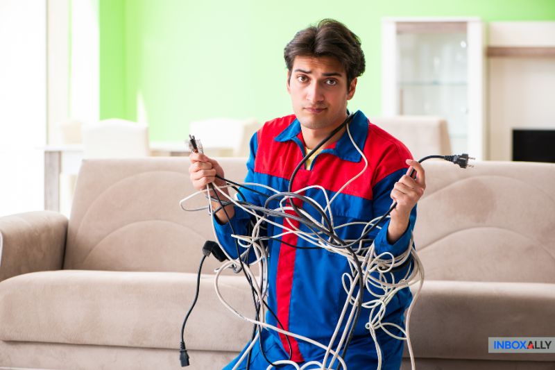 Um homem em um uniforme azul e vermelho segura um feixe emaranhado de cabos elétricos, em uma sala de estar com um sofá bege - como solucionar problemas de entrega de e-mail ou resolver um erro de FPS.