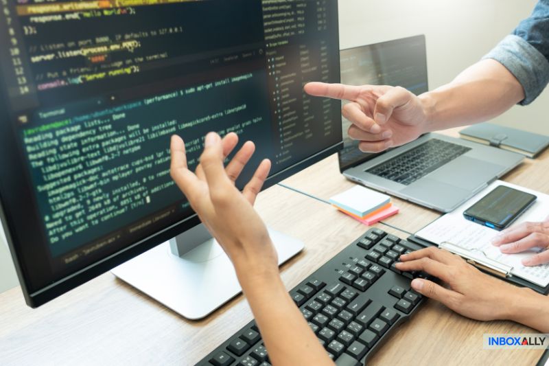 Duas pessoas discutem um erro de SPF exibido em um grande monitor de computador em uma mesa com teclado, laptop, smartphone e blocos de notas enquanto solucionam problemas de entrega de e-mail.