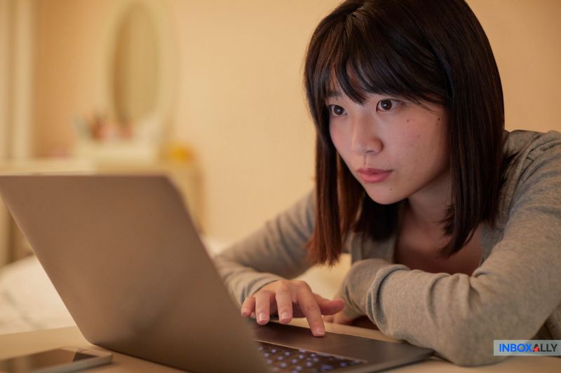 Uma mulher deita-se em uma cama e olha atentamente para a tela de um laptop enquanto digita, pesquisando o SPF Permerror e seu impacto na capacidade de entrega de e-mails, em um quarto com iluminação suave.