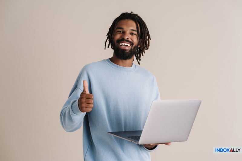 Um homem com dreadlocks vestindo um suéter azul claro segura um laptop em uma das mãos e faz sinal de positivo com a outra, em um fundo bege simples - destacando o sucesso na capacidade de entrega de e-mails, apesar de enfrentar um erro de SPF.
