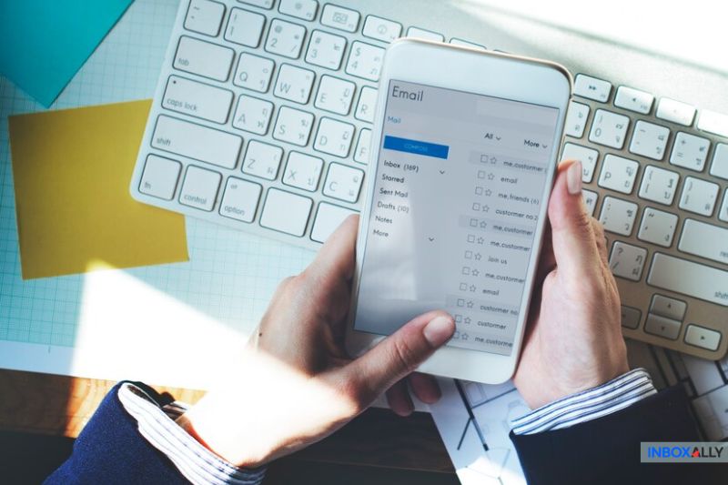 A person holds a smartphone displaying an email inbox with checkboxes, navigating through WooCommerce notifications to prevent spam, in front of a keyboard and laptop.