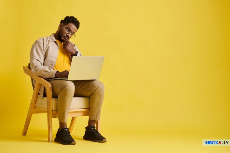 A man sits on a wooden chair against a yellow background, thoughtfully examining his laptop screen, perhaps contemplating the setup of his WooCommerce store.