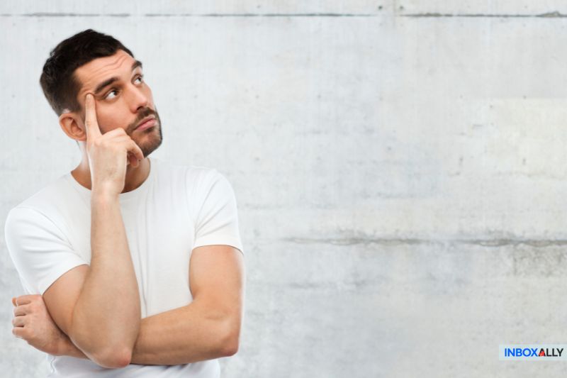 A man in a white T-shirt looks up thoughtfully, touching his temple with one finger, as though contemplating the complexities of WooCommerce, against a concrete wall background.