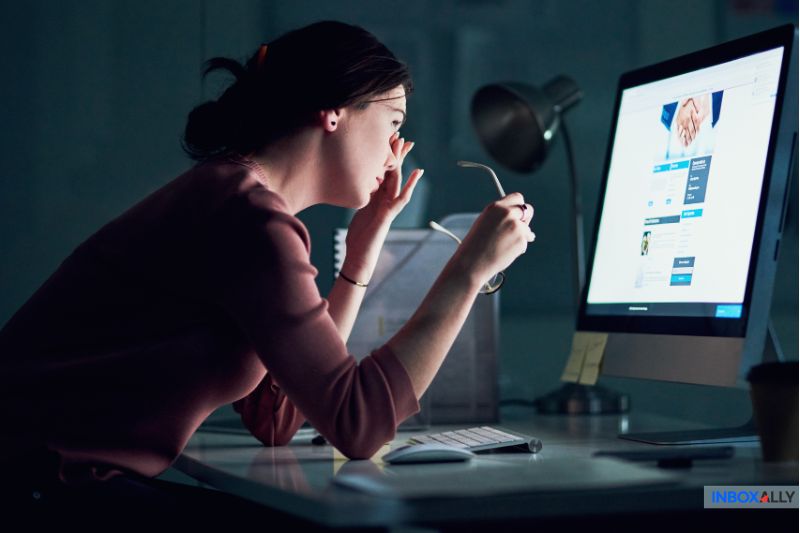 A woman sits at a desk, holding her glasses and looking closely at a computer screen displaying an email error—possibly a 550 5.7.0 Local Policy Violation—with a lamp and office supplies nearby.