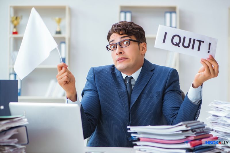 A man in a suit sits at a cluttered desk, holding a white flag and a sign reading "I QUIT!"—his resigned expression says it all, as if overwhelmed by endless email errors or an SMTP error like "550 5.7.0 Local Policy Violation.